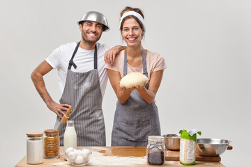 Indoor shot of positive friendly hard working woman and man chefs or cooks prepared fresh dough for baking pastry, being in high spirit, prepare festive dinner for guests, spend free time at kitchen