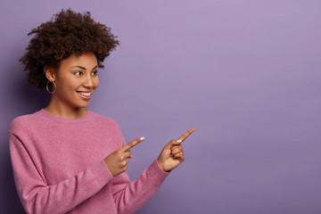 Beautiful curly ethnic woman points at copy space, suggests click link or follow page to find out...