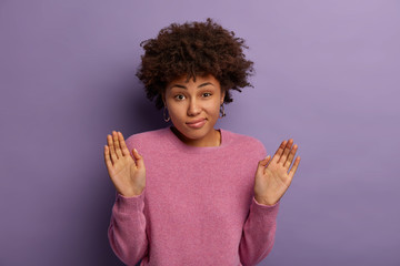 What you want from me, who cares. Unsure indifferent dark skinned woman raises palms, has hesitant expression, not gonna help, stands confused and perplexed indoor, isolated on purple background