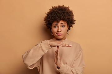 Photo of puzzled dark skinned woma makes time out gesture, has serious frustrated expression, curly...