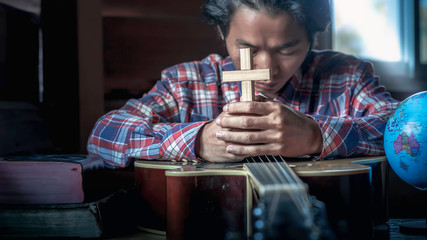 Asian man holding the crucifix and praying on acoustic guitar at home. Christian worship concept.