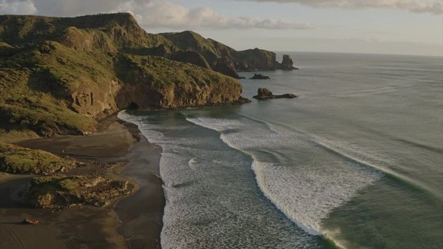 Aerial flying over calm West Coast ocean with waves crashing onto black sand rocky beach during West Coast sunset. Te Henga, Bethells Beach, Auckland, New Zealand.