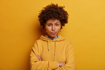 Fototapeta na wymiar Skeptic angry ethnic woman expresses suspision, stands with arms folded, pouts lips and waits for explanations, being angry with someone, wears casual clothes, isolated on yellow studio wall