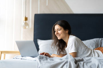 Beautiful young brunette girl with a laptop lying on the bed. Stylish modern interior. A cozy workplace. Shopping on the Internet.