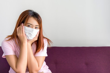 portrait of young Asian woman sitting on sofa at living room in her house and wearing face medical mask. Flu epidemic, dust allergy, protection against virus. Covid-19, corona virus pandemic