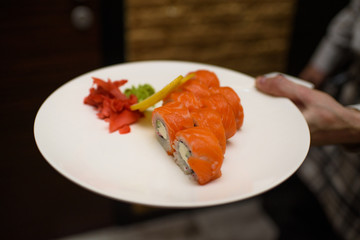 The waiter holds a white plate on which are rolls Philadelphia, wasabi, ginger and a lemon. Serving for guests. Asian food.