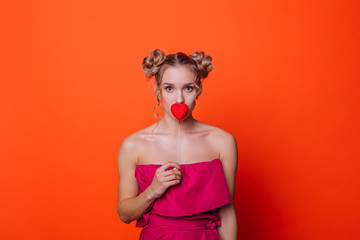 The girl in a red dress on a orange background in the studio. Blonde girl with two hair knots holding red heart on the stick, looking to the camera and smiling.