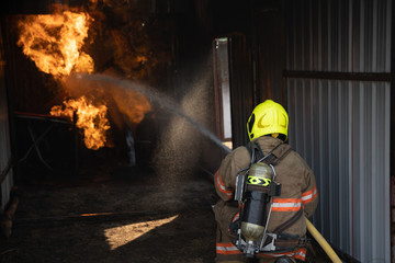 Fototapeta premium Rear view of Fireman wear fire protection suit walking into the fire training room and Spray water Fire at fire station. Training fire drill Concept.