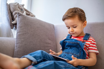 Cute boy using a mobile phone. A little boy looking to smartphone lying on the couch at home. Toddler boy using tablet or smartphone. Cute three years old boy sitting at home using digital device.