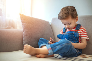 Child and electronic devices concept. Little boy sitting at home and playing with mobile phone. Kids and mobile devices. Children and technology. Fun time with mobile phone for kids.