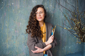 Portrait of beautiful girl with curly hair and glasses posing with brushes for watercolor, creative workplace or studio