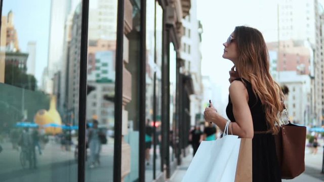 Young beautiful woman with bags standing near the shop-window and using the smartphone. Slow motion.