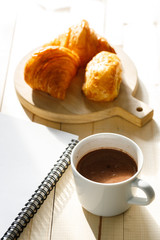 A cup of hot chocolate and croissant on wooden background with notebook.  Work from home.