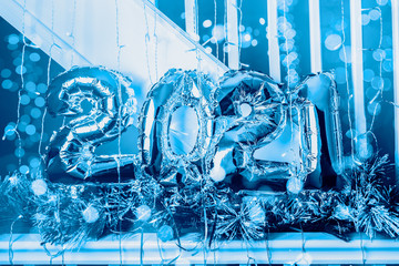 Balloon Bunting for celebration of New Year 2021 made from Silver Number Balloons. Holiday Party Decoration or postcard concept with xmas lights and christmas tree branches. Toned in blue