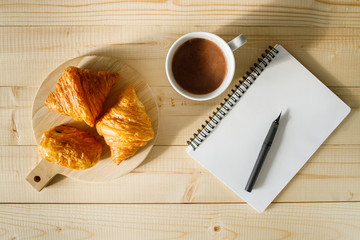 A cup of hot chocolate and croissant on wooden background with notebook.  Work from home.