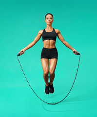 Sporty girl exercising with skipping rope. Photo of girl in black sportswear on turquoise background. Dynamic movement. Full length. Sports and healthy lifestyle - obrazy, fototapety, plakaty