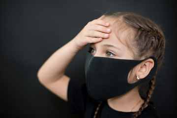 Young girl in protective sterile medical mask on her face has heat temperature, hold head with hand on a black background. Feeling sick, bad, suffering. Pandemic coronavirus symptom.