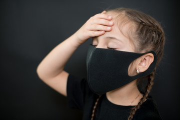 Young girl in protective sterile medical mask on her face has heat temperature, hold head with hand on a black background. Feeling sick, bad, suffering. Pandemic coronavirus symptom.