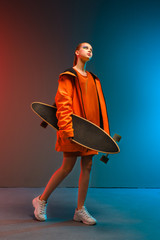 A young girl with bright makeup holds a longboard in her hands on a fashionable neon color background. Portrait of a girl skater