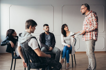 Handsome senior professional therapist stands in front of a group of people of different ages...