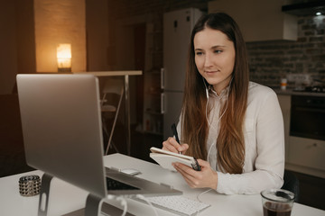 Remote work. A caucasian woman with headphones working remotely on her laptop. A girl in a white shirt doing notes during an online business briefing at her cozy home workplace.