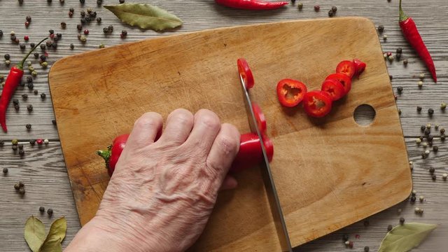 Using Knife To Cutting Ramiro Red Hot Pepper On Cutting Wooden Board. Top View. Close Up. Green Bay Leaf, Multi Color Peppercorns And Bright Red Chili Pepper On Gray Wooden Background..