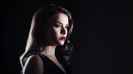 Fototapeta premium Portrait of a young luxurious woman in a low key. Bright makeup with scarlet lipstick, hairstyle with curls,
