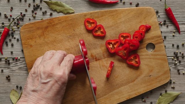 Using Knife To Cutting Ramiro Red Hot Pepper On Cutting Wooden Board. Top View. Close Up. Green Bay Leaf, Multi Color Peppercorns And Bright Red Chili Pepper On Gray Wooden Background..