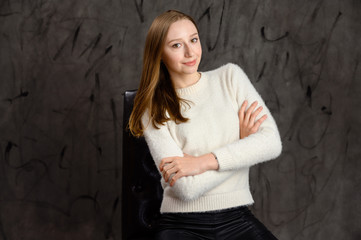 Studio portrait of a pretty happy blonde girl. Model posing on a chair on a gray stylish background.