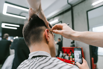 Barber shaves a straight razor, the guy sits in the barber shop's wing and takes a photo on a smartphone in the mirror.Male hairdresser creates a stylish hairstyle for the client,uses a straight razor