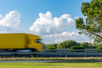 Yellow truck in motion driving on the highway next to a tree and a blue sky with clouds.