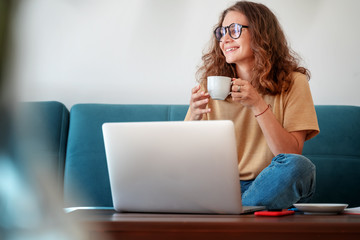 Smiling happy curly young woman in eyeglasses enjoying morning coffee using laptop sitting at home...