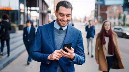 Caucasian Businessman in a Suit is Using a Smartphone on a Street in Downtown. Other Office People Walk Past. He Smiles and Looks Successful. He's Browsing the Web on his Device.
