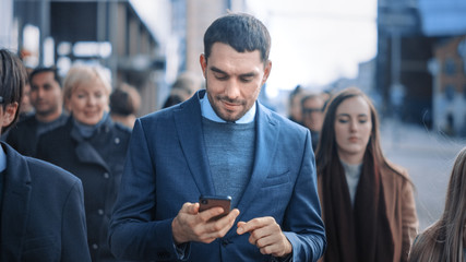 Caucasian Businessman in a Suit is Using a Smartphone on a Street in Downtown. Other Office People Commute in a Crowd. He's Confident and Looks Successful. He's Browsing the Web on his Device.