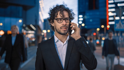 Young Stylish Businessman with Curly Hair is Talking on a Phone While Walking in the City. He's Wearing Glasses. Office People Walk By. It's Evening with Atmospheric Urban Lights in the Background.