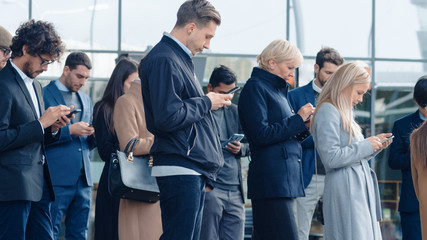 Obraz premium Office Managers and Businessmen are Standing in Front of a Modern Glass Office Building and Use Their Smartphones. People are Dressed Smartly and Look Successful. They are Occupied by Their Devices.