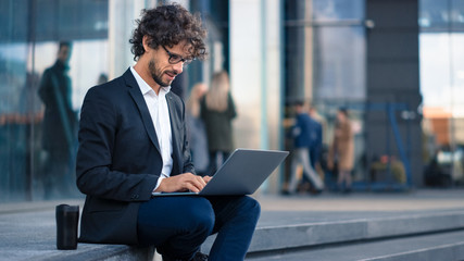 Handsome Businessman in a Suit is Sitting on Steps next to Business Center and Working on a Laptop on a Street in a City. Office People Walk By to Work.