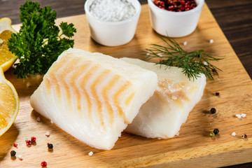Fresh raw cod with seasonings and vegetables served on cutting board on wooden table
