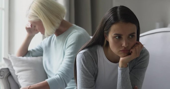 Head shot depressed two generations women sitting back to back, ignoring each other after fight. Depressed middle aged mother and young selfish stubborn grownup daughter feeling stressed after argue.