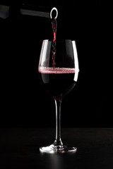 Naklejka na ściany i meble Red wine being poured into a glass and splashing over the side. Low key black background.