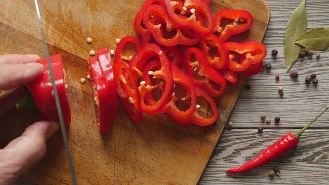 Using Knife To Cutting Ramiro Red Hot Pepper On Cutting Wooden Board. Top View. Close Up. Green Bay Leaf, Multi Color Peppercorns And Bright Red Chili Pepper On Gray Wooden Background..