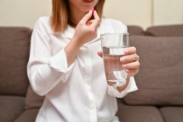 Close up Asian woman holding pill and a glass of water sitting on sofa. Concept of taking daily medicine, Multivitamins and supplements and medicine to cure head ache, stomach pain sedation meds.