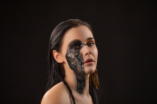Portrait of young sensual brunette woman with black raven bird body art on her face.