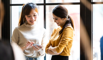Two Charming Asian business women with the smartphone standing on a window. Girl shows her friend image on screen of smartphone. Young people problems with cell phone addiction
