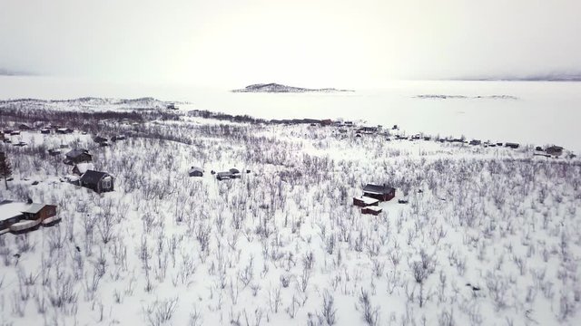 Wooden houses by the beautiful white frozen lake of the peaceful village of Abisko, Sweden - aerial