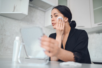 Obraz premium Beautiful brunette woman removing makeup from her face with mirror. Cute beautiful natural beauty girl cleaning face with cotton sponge. Cosmetology and spa, problem skin care, acne treatment concept