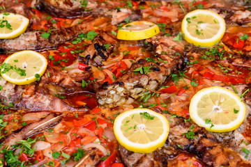 Close up of large portion of cooked fish with red peppers, onion, parsley and fresh yellow lemons in a large pan at a street food festival, ready to eat healthy seafood
