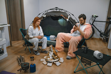 Couple sitting in folding chairs by the tent and bonfire. Camping in the apartment. Adventures at home, traveling around the room. quarantine entertainment. Use smartphone. compact table