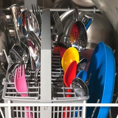 Stainless steel spoons and forks and children's multi-colored plastic dishes in a cutlery basket inside the dishwasher