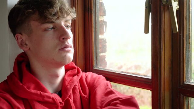 Sad teen male young man boy teenager wearing a red hoodie sitting looking out of a window during Coronavirus COVID-19 pandemic self isolation lockdown quarantine